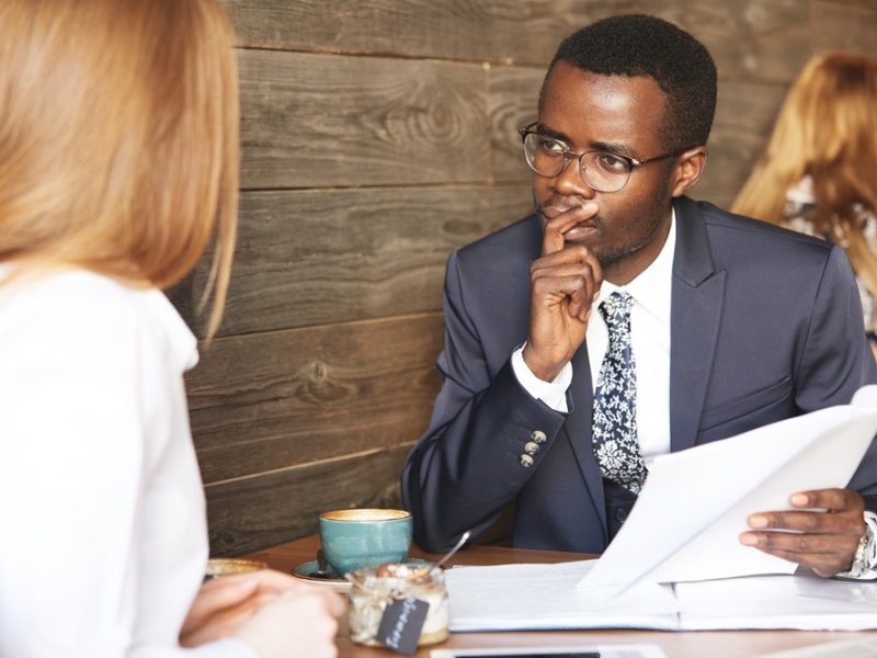 Recruiting and human resource concept. African HR director in suit conducting an interview, looking at female Caucasian candidate with pensive concentrated expression, holding finger on his chin